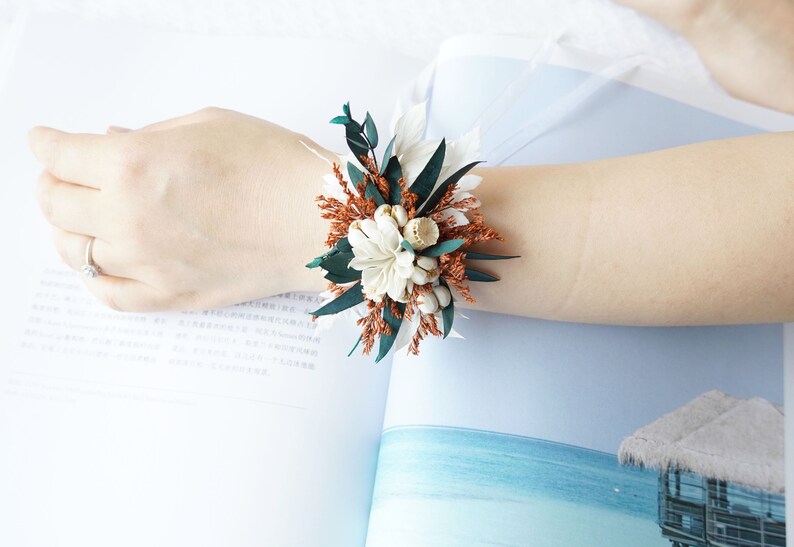May include: A wrist corsage made with dried flowers and greenery. The corsage features white flowers, brown dried grasses, and green leaves.