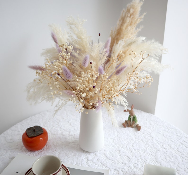 Pu&ograve; includere: Un vaso di ceramica bianco contiene una composizione floreale secca. La composizione comprende soffice erba della pampa beige, delicato velo da sposa bianco e morbida erba coda di coniglio lavanda. Un frutto arancione e una statuina di coniglio in ceramica sono sul tavolo.