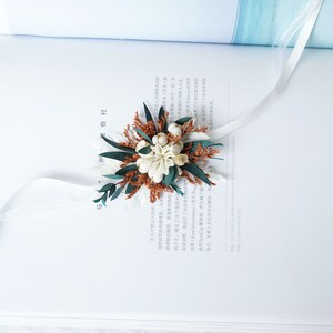 May include: A white and brown dried flower bouquet with green leaves and white ribbon. The bouquet is tied with a white ribbon and is sitting on a white book.