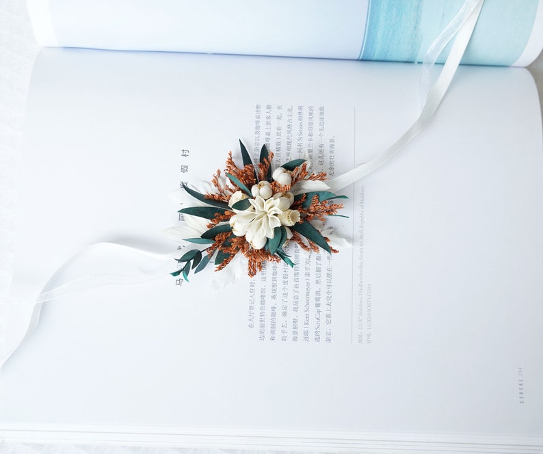 May include: A white and brown dried flower bouquet with green leaves and white ribbon. The bouquet is tied with a white ribbon and is sitting on a white book.