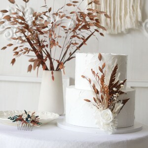 May include: A two-tiered white wedding cake decorated with dried flowers and greenery. The cake is on a white cake stand and is surrounded by a white tablecloth.