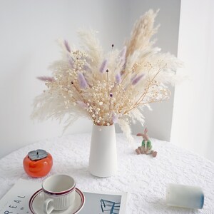 Pu&ograve; includere: Un vaso di ceramica bianco contiene un bouquet di fiori secchi, tra cui erba della pampa, rametti color lavanda e piccoli fiori bianchi. Una tazza e un piattino, un caco in ceramica e una piccola statuina di coniglio sono su una tovaglia di pizzo bianco.