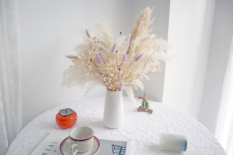 Pu&ograve; includere: Un vaso di ceramica bianco contiene un bouquet di fiori secchi, tra cui erba della pampa, rametti color lavanda e piccoli fiori bianchi. Una tazza e un piattino, un caco in ceramica e una piccola statuina di coniglio sono su una tovaglia di pizzo bianco.