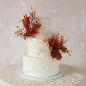 May include: A three-tiered white cake decorated with dried flowers and roses in shades of orange and red. The cake sits on a white cake stand and is displayed on a white tablecloth.