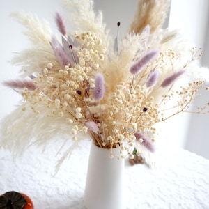 Pu&ograve; includere: Un vaso di ceramica bianco contiene una composizione floreale secca. La composizione presenta soffice erba della pampa color crema, delicato velo da sposa bianco e morbida erba coda di coniglio lavanda. Il bouquet ha una palette di colori neutri.