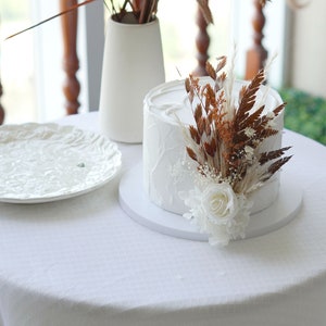 May include: A white cake with a textured buttercream frosting is decorated with dried flowers and a single white rose. The cake is on a white cake stand and a white tablecloth.