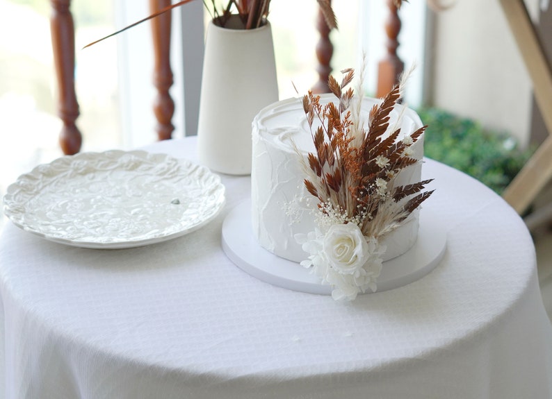 May include: A white cake with a textured buttercream frosting is decorated with dried flowers and a single white rose. The cake is on a white cake stand and a white tablecloth.