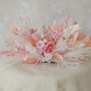 May include: A pink and white dried flower arrangement with fluffy white pampas grass, pink roses, and other dried flowers.