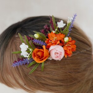 May include: A floral hair clip featuring orange roses, pink and orange blossoms, white flowers, and purple sprigs. The arrangement is set against brown hair, creating a vibrant contrast. A decorative hair accessory.