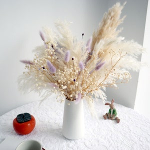 Pu&ograve; includere: Un vaso di ceramica bianco pieno di una composizione floreale secca. La composizione include soffice erba della pampa beige, delicato gypsophila bianco e erba coda di coniglio lavanda. Un caco in ceramica e una piccola statuina di coniglio sono sul tavolo.