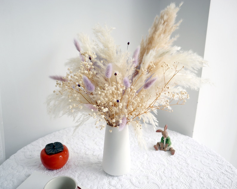 Pu&ograve; includere: Un vaso di ceramica bianco pieno di una composizione floreale secca. La composizione include soffice erba della pampa beige, delicato gypsophila bianco e erba coda di coniglio lavanda. Un caco in ceramica e una piccola statuina di coniglio sono sul tavolo.