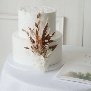 May include: A two-tiered white wedding cake decorated with dried flowers and a single white rose. The cake is on a white cake stand and a magazine with the word "Cereal" printed on the cover is in the foreground.