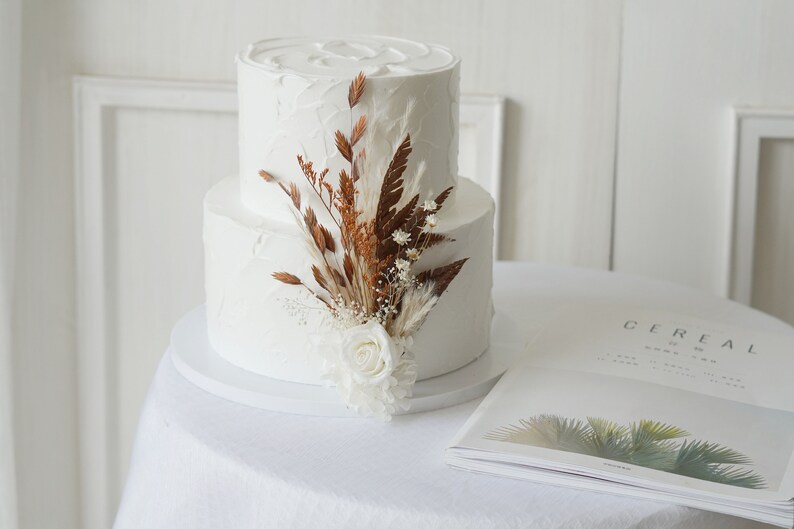 May include: A two-tiered white wedding cake decorated with dried flowers and a single white rose. The cake is on a white cake stand and a magazine with the word "Cereal" printed on the cover is in the foreground.