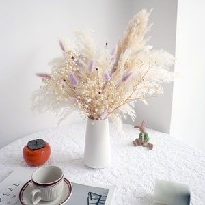 Pu&ograve; includere: Un vaso di ceramica bianco contiene un bouquet di fiori secchi. L'arrangiamento comprende erba della pampa color crema, delicato velo da sposa bianco e morbidi accenti viola. Una tazza e un piattino, un caco in ceramica e una piccola statuina di coniglio sono sul tavolo.