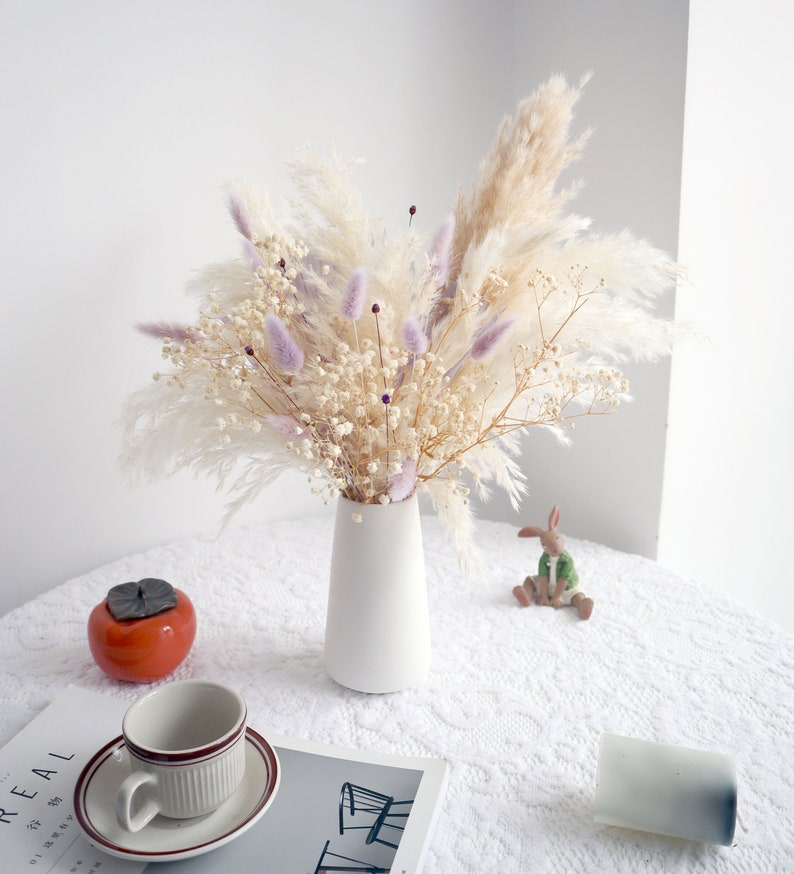 Pu&ograve; includere: Un vaso di ceramica bianco contiene un bouquet di fiori secchi. L'arrangiamento comprende erba della pampa color crema, delicato velo da sposa bianco e morbidi accenti viola. Una tazza e un piattino, un caco in ceramica e una piccola statuina di coniglio sono sul tavolo.