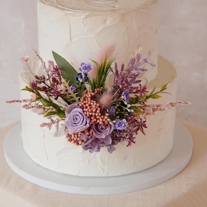 Puede incluir: Un pastel blanco de dos niveles con glaseado texturizado, adornado con un arreglo floral de rosas lavanda, flores silvestres moradas y vegetación. El pastel está sobre un soporte blanco, perfecto para una boda o celebración.