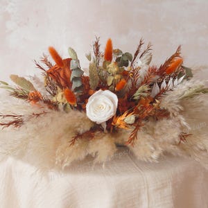 May include: A dried floral arrangement featuring a large white rose at the center, surrounded by orange, brown, and beige dried flowers and pampas grass. The arrangement is set on a cream-colored surface.