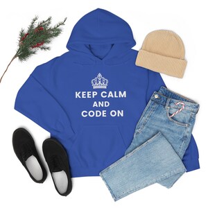 May include: A royal blue hoodie with a white crown and the text "Keep Calm and Code On". The hoodie is laid out on a white surface with a pair of black shoes, a pair of blue jeans, and a beige beanie.
