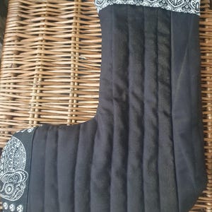May include: Black quilted Christmas stocking with a white and black paisley patterned cuff and toe. The stocking has vertical stitched lines and is made of a soft fabric. The cuff and toe have a decorative design.