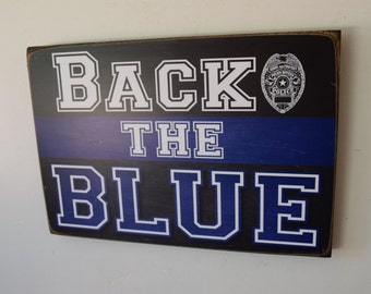 May include: A wooden sign with the text "Back the Blue" in white letters with a black outline. The sign also features a blue and black thin blue line and a silver police badge.