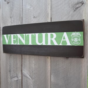 May include: A wooden sign with the word "VENTURA" in white letters on a green background. The sign also features a black and white emblem with the letters "U.S." and a bird.