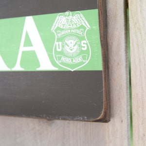 May include: A wooden sign with a green stripe and the words "U.S. BORDER PATROL PATROL AGENT" in white. The sign is on a wooden background.