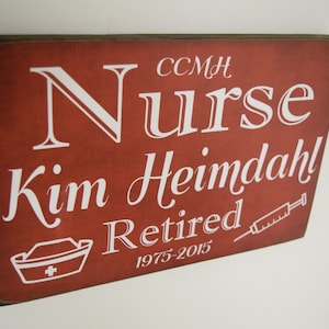 Puede incluir: Un letrero de madera rojo con texto blanco que dice "Nurse Kim Heimdahl Retired 1975-2015". El letrero también tiene una pequeña imagen blanca de un gorro de enfermera y una jeringa.