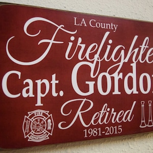 Puede incluir: Un letrero de madera rojo con texto blanco que dice "LA County Firefighter Capt. Gordon Retired 1981-2015". El letrero tambi&eacute;n presenta un logotipo del departamento de bomberos y dos trompetas cruzadas.