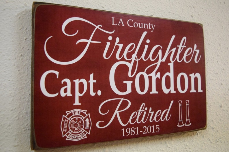 Puede incluir: Un letrero de madera rojo con texto blanco que dice "LA County Firefighter Capt. Gordon Retired 1981-2015". El letrero tambi&eacute;n presenta un logotipo del departamento de bomberos y dos trompetas cruzadas.