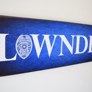 K&ouml;nnte beinhalten: Ein blaues Holzschild mit dem Wort "LOWNDES" in wei&szlig;en Buchstaben. Ein wei&szlig;es Polizeibadge mit einem Stern und den Worten "Hollywood, Florida Police Dept." befindet sich auf der linken Seite des Schildes.