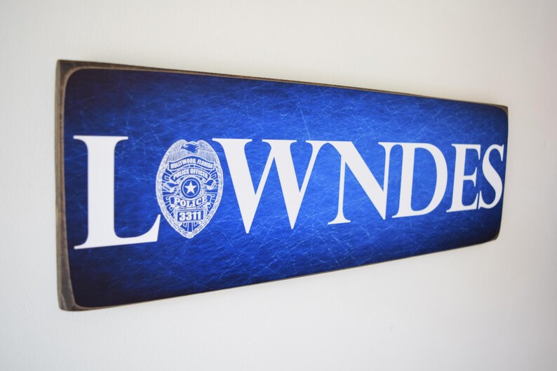 K&ouml;nnte beinhalten: Ein blaues Holzschild mit dem Wort "LOWNDES" in wei&szlig;en Buchstaben. Ein wei&szlig;es Polizeibadge mit einem Stern und den Worten "Hollywood, Florida Police Dept." befindet sich auf der linken Seite des Schildes.