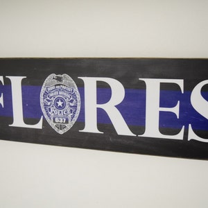 Puede incluir: Un letrero de madera con fondo negro y azul y una línea blanca fina. El letrero tiene el nombre "FLORES" en letras blancas y una placa de policía con el texto "Serve and Protect Police Officer" y "Police 637".