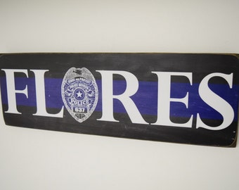 May include: A wooden sign with a black and blue background and a white thin line. The sign has the name "FLORES" in white letters and a police badge with the text "Serve and Protect Police Officer" and "Police 637".