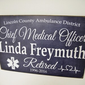 Puede incluir: Un letrero de madera azul oscuro con texto blanco que dice "Lincoln County Ambulance District Chief Medical Officer Linda Freymuth Retired 1996-2016". El letrero también presenta un símbolo blanco de estrella de la vida y un gráfico blanco de latido del corazón.