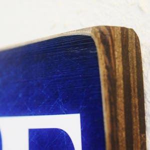 K&ouml;nnte beinhalten: Nahaufnahme eines blauen Holzschildes mit wei&szlig;en Buchstaben. Das Schild hat einen abgenutzten Look und ist aus Sperrholz gefertigt.