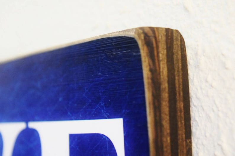 K&ouml;nnte beinhalten: Nahaufnahme eines blauen Holzschildes mit wei&szlig;en Buchstaben. Das Schild hat einen abgenutzten Look und ist aus Sperrholz gefertigt.