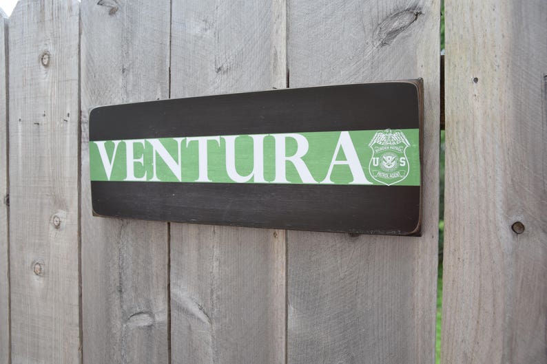 May include: A wooden sign with the word "VENTURA" in white letters on a green background. The sign also features a black and white emblem with the letters "U.S." and a bird.