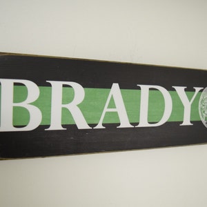 May include: A wooden sign with a black background and a green stripe. The name "BRADY" is written in white letters on the sign. A thin green line with a police badge is on the right side of the sign.