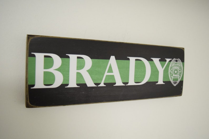 May include: A wooden sign with a black background and a green stripe. The name "BRADY" is written in white letters on the sign. A thin green line with a police badge is on the right side of the sign.