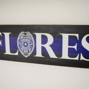 K&ouml;nnte beinhalten: Ein Holzschild mit schwarzem und blauem Hintergrund und dem Namen "FLORES" in wei&szlig;en Buchstaben. Ein silbernes Polizeibadge mit einem blauen Band ist mittig auf dem Schild angebracht.