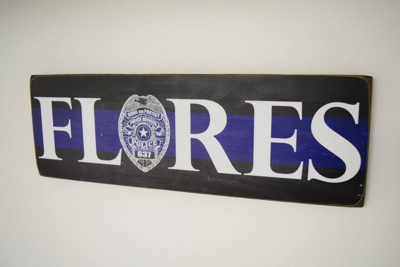 K&ouml;nnte beinhalten: Ein Holzschild mit schwarzem und blauem Hintergrund und dem Namen "FLORES" in wei&szlig;en Buchstaben. Ein silbernes Polizeibadge mit einem blauen Band ist mittig auf dem Schild angebracht.