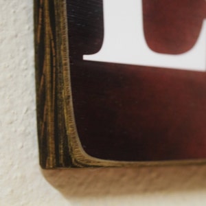 Puede incluir: Primer plano del borde de un letrero de madera con un marco de madera marr&oacute;n oscuro. El letrero tiene un fondo rojo con letras blancas.