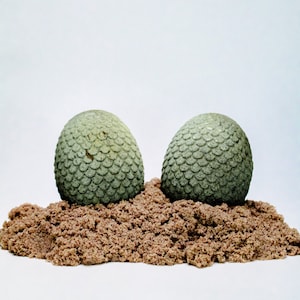 May include: Two green dragon eggs with a scaly texture are partially buried in brown sand.