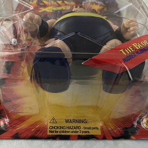 May include: A plastic package containing a black rubber toy figure of The Blob, a character from the movie The Blob. The figure is in a sitting position with its arms outstretched. The package has a red and yellow background with the words "The Blob Rubber Blubber Ball" printed on it. A warning label is visible on the bottom of the package.