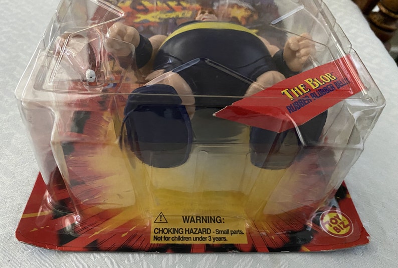 May include: A plastic package containing a black rubber toy figure of The Blob, a character from the movie The Blob. The figure is in a sitting position with its arms outstretched. The package has a red and yellow background with the words "The Blob Rubber Blubber Ball" printed on it. A warning label is visible on the bottom of the package.