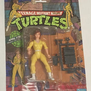 May include: Teenage Mutant Ninja Turtles action figure of April O'Neil in a yellow jumpsuit with a gun and a stand. The packaging features the Teenage Mutant Ninja Turtles logo and the text "Amazing Arm Movement! Deluxe Weapons Assortment Included!"