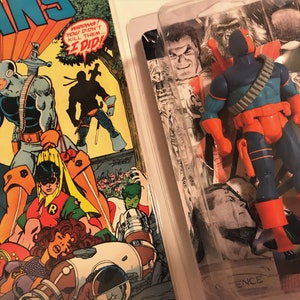 以下が含まれることがあります： プラスチック製のブリスターパックに入った、DCコミックスのスーパーヴィラン、デスストロークの青とオレンジのフィギュア。フィギュアは片手に剣、もう片方に銃を持ってポーズをとっている。ブリスターパックの背景には、他のDCコミックスのキャラクターの白黒画像のコラージュが描かれている。