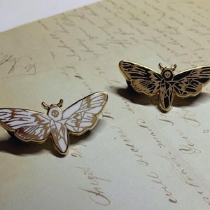 May include: Two gold-tone moth pins, one with a white moth and one with a black moth. Both moths have a skull and crossbones design on their bodies.