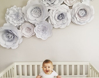 May include: A white crib with a baby in it, surrounded by a wall decorated with large, white paper flowers.