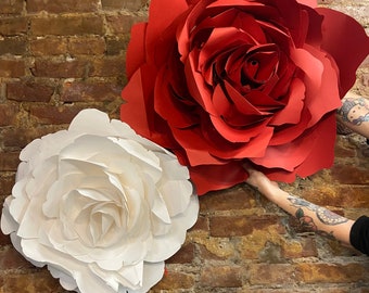 May include: Two large paper roses, one red and one white, are displayed against a brick wall. The roses are made of paper and have a layered, three-dimensional appearance.